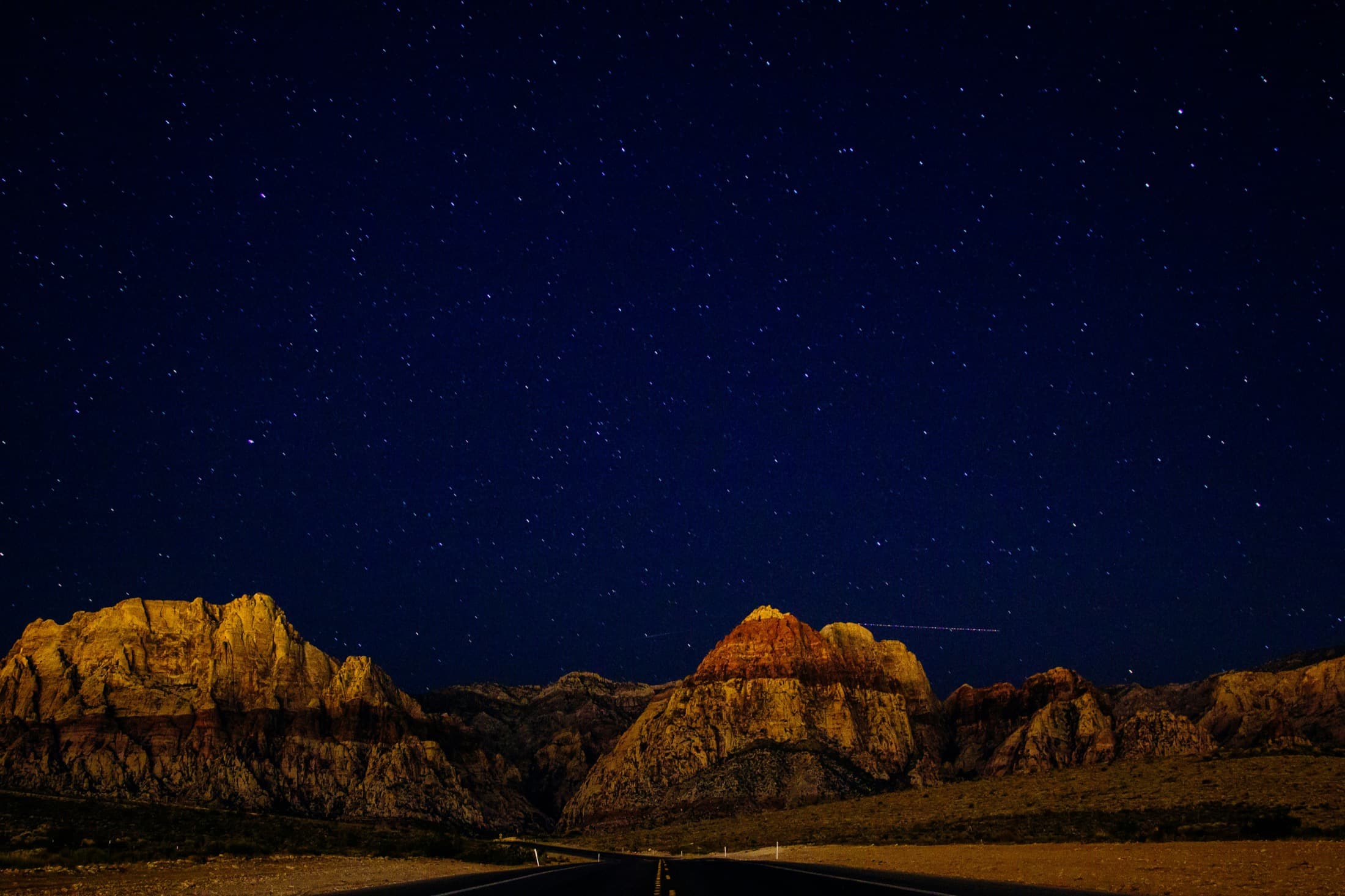 An empty desert road running toward red rock under a starlit sky