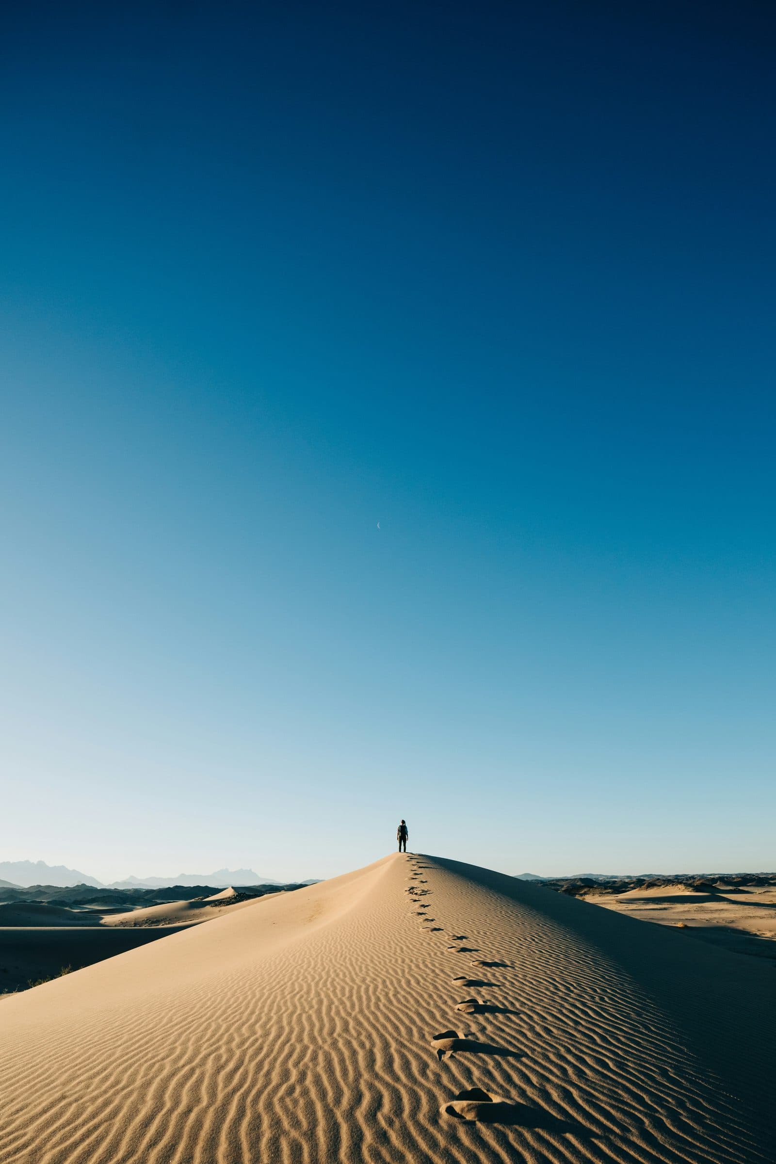 A single set of footprints running up the ridge of a dune toward a man near the top.